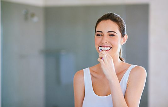 Woman in white shirt smiling while brushing her teeth